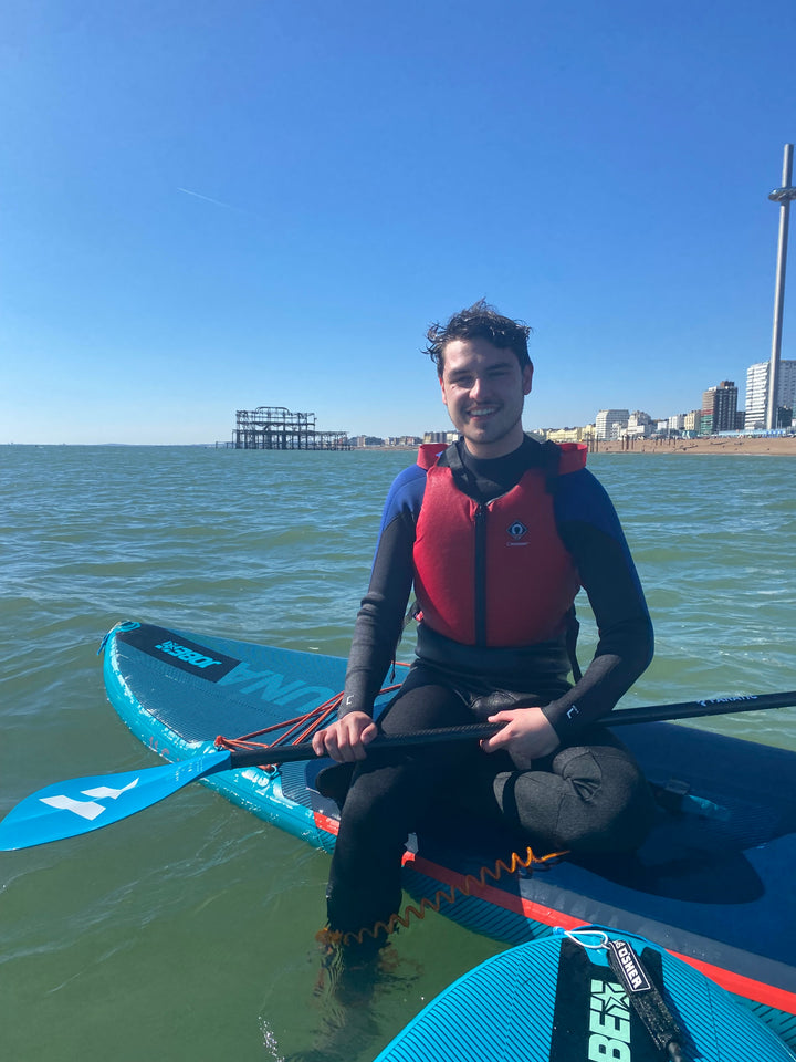 A person sitting on a paddleboard in the water, wearing a life jacket and holding a paddle, with Brighton Beach and a pier in the background.