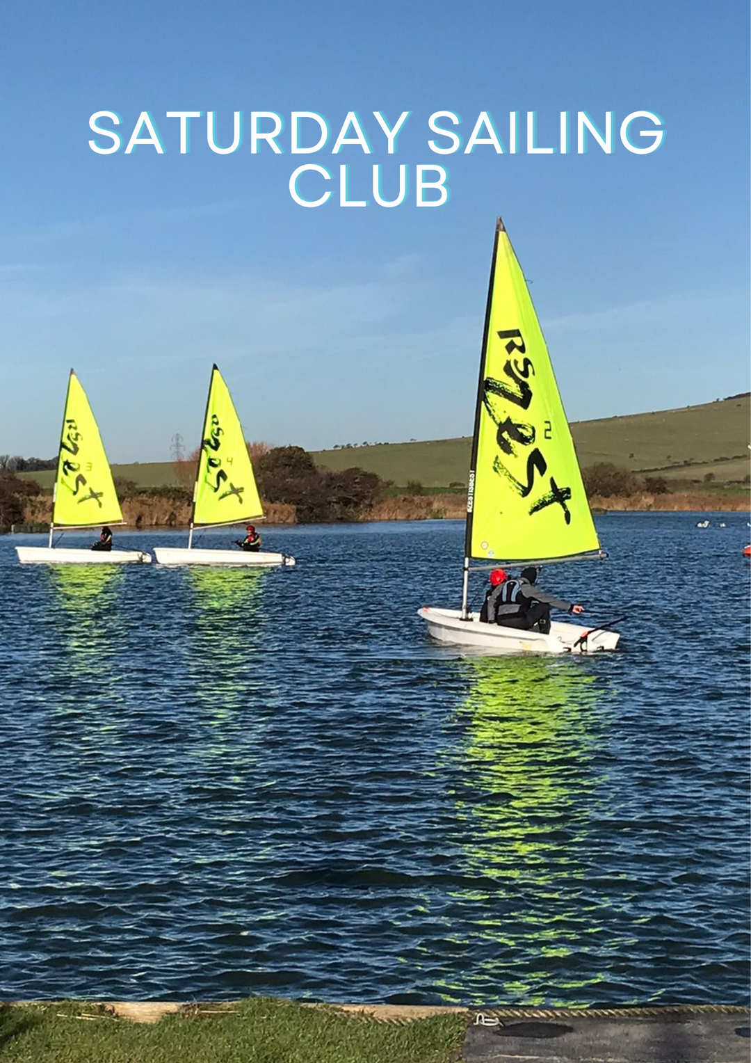 Two sailboats with bright yellow sails marked 'SATURDAY SAILING CLUB', sailing on a calm lake on a clear day.